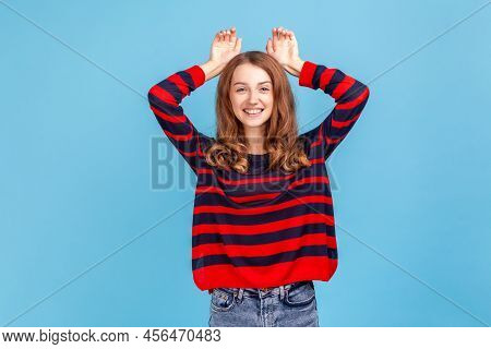 Portrait Of Cheerful Woman Wearing Striped Casual Style Sweater Showing Bunny Ears Gesture And Looki