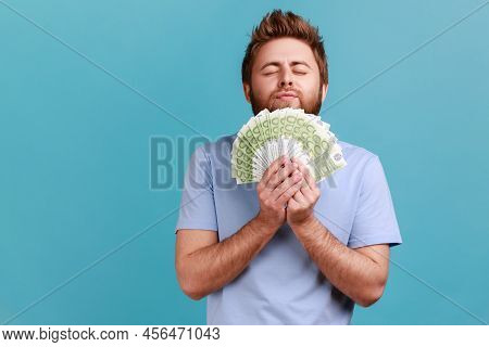 Portrait Of Satisfied Bearded Man Smelling Earned Euro Banknotes, Enjoying Success And Big Profit, W