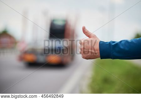 Close Up Of Mans Hand Hitchhiking By Roadside. Male Hand Showing Thumbs Up Gesture Outdoors On Blurr