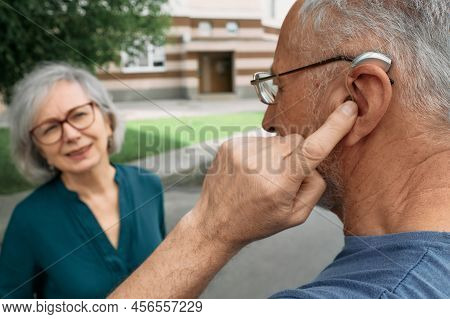 Mature Man With A Hearing Impairment Uses A Hearing Aid To Communicate With His Female Senior Friend