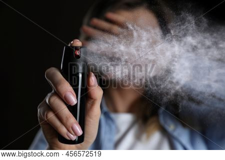 Young Woman Covering Eyes With Hand And Using Pepper Spray On Black Background