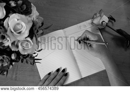 Female Hands Writing In Open Notebook And Bouquet Of Roses On Old Wooden Table. Planning Notebook Wi