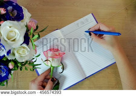Female Hands Writing In Open Notebook And Bouquet Of Roses On Old Wooden Table. Planning Notebook Wi