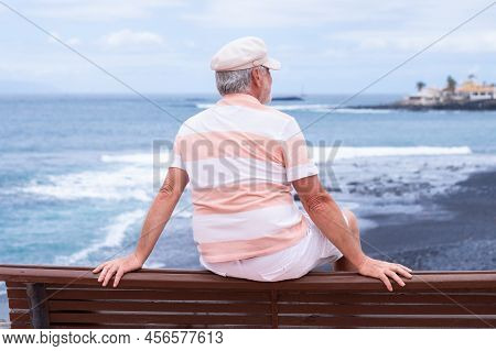 Rear View Of Senior Man With Beard And Hat Sitting Outdoors At Sea Looking At Horizon Enjoying Freed