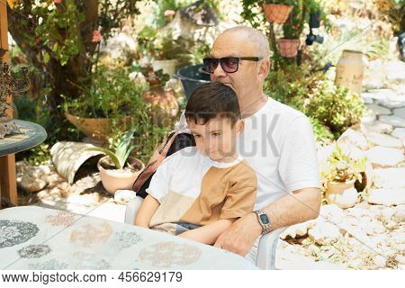 Little Boy Sits On His Grandfathers Lap As They Both Rest In The Backyard. Happy Grandchild With Gra