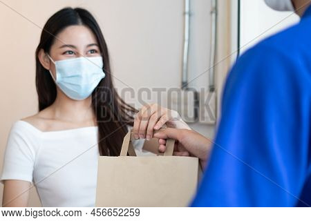 Smiling Young Asian Woman,client Accepting A Delivery Of Bag, Cardboard Box From Delivery Man Wearin
