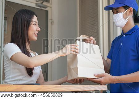 Smiling Young Asian Woman,client Accepting A Delivery Of Bag, Cardboard Box From Delivery Man Wearin