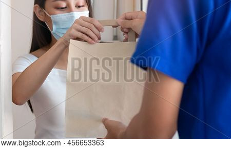 Smiling Young Asian Woman,client Accepting A Delivery Of Bag, Cardboard Box From Delivery Man Wearin