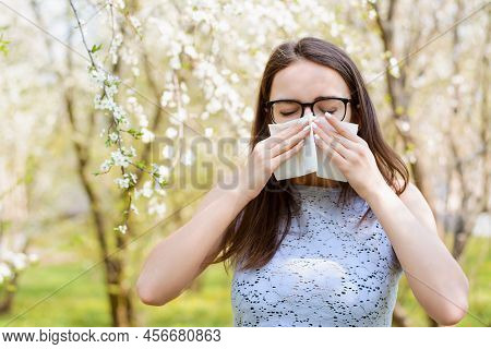 Allergic Woman Blowing Her Nose To The White Handkerchief Against Blooming Trees In Park In Spring. 