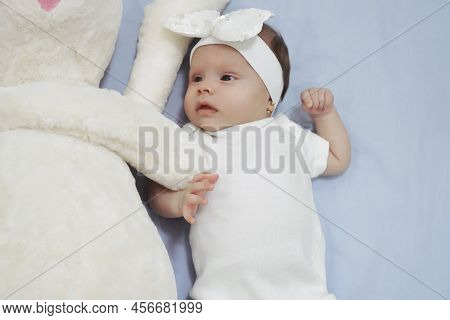 White Dreesed Newborn Baby With Her White Bunny Toy. Cute Mixed Race Baby Girl Looking Into The Came