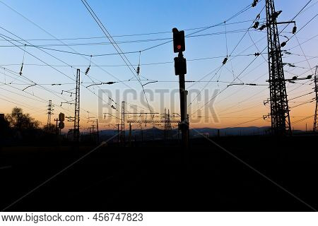 Red Signal Semaphore On The Railroad On A Dark Background At Dusk. Black Silhouettes Catenary And Ma