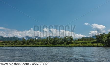 A Calm Blue River Flows Along The Shore. Lush Green Vegetation And A Picturesque Mountain Range Agai
