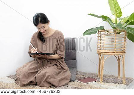 Asian woman sits in her living room writing a diary or notebook on the carpet, Weekends or holidays 