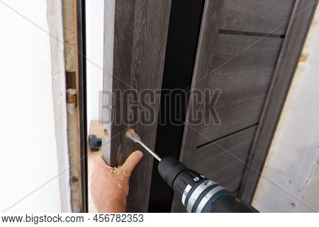 A Carpenter Is Installing A Doorknob With A Latch On A Wooden Door. Close-up Of A Hand With A Tool. 