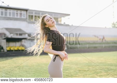 Girl In Sportswear Throws Her Head Back Against The Background Of The Stadium. A Woman Stands And Ba