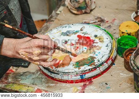 The Artist Holds A Brush In His Hand Near A Palette With Bright Colors In An Art Workshop