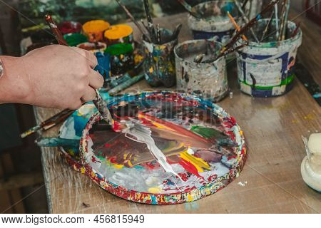 The Artist Holds A Brush In His Hand Near A Palette With Bright Colors In An Art Workshop