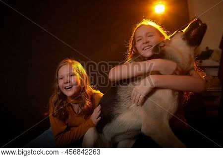 Two Cheerful Joyfull Sisters Having Fun Together And With Big German Shepherd Dog In Dark Studio Wit