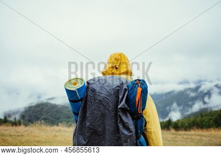 The Back Of A Male Tourist In A Raincoat Stands In The Rain On In The Mountains On The Background Of