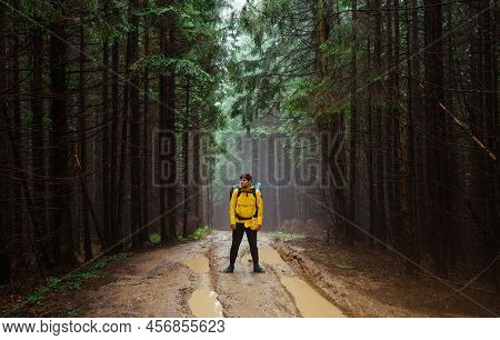Male Hiker Stands On A Winding Road In The Mountains With A Backpack On His Back Against The Backdro
