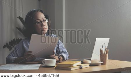 Seriously Senior Woman Tworking At Home, Looking At Laptop And Waving Paper Correspondence, Sitting 
