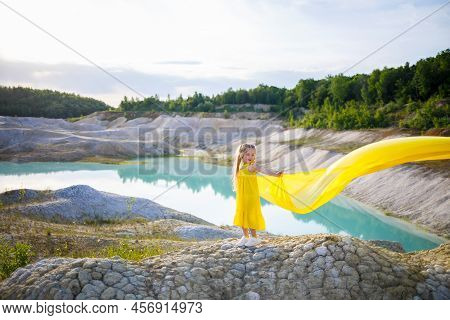 A Small 6-7 Years Old Ukrainian Girl In A Yellow Dress With Wings In Yellow Fabric By The Lake. Blue