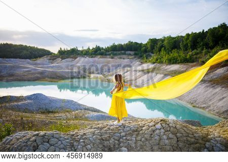 A Small 6-7 Years Old Ukrainian Girl In A Yellow Dress With Wings In Yellow Fabric By The Lake. Blue