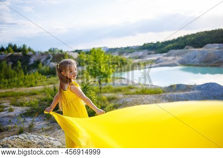 A Small 6-7 Years Old Ukrainian Girl In A Yellow Dress With Wings In Yellow Fabric By The Lake. Blue