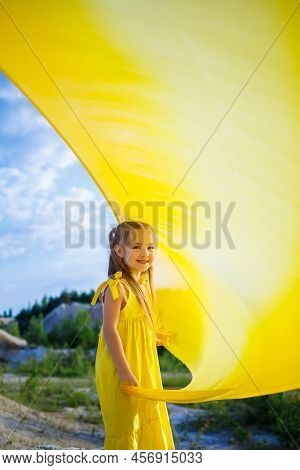 A Small 6-7 Years Old Ukrainian Girl In A Yellow Dress With Wings In Yellow Fabric By The Lake. Blue