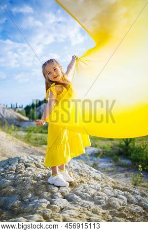 A Small 6-7 Years Old Ukrainian Girl In A Yellow Dress With Wings In Yellow Fabric By The Lake. Blue