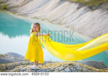A Small 6-7 Years Old Ukrainian Girl In A Yellow Dress With Wings In Yellow Fabric By The Lake. Blue