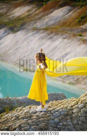 A Small 6-7 Years Old Ukrainian Girl In A Yellow Dress With Wings In Yellow Fabric By The Lake. Blue
