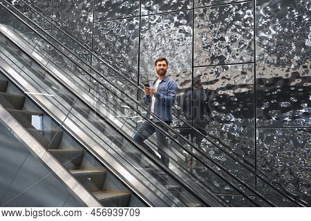 Smiling Bearded Male Using Mobile Phone, While Lifting On Escalator. Side View Of Happy Man Standing