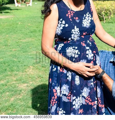 Indian Couple Posing For Maternity Baby Shoot. The Couple Is Posing In A Lawn With Green Grass And T