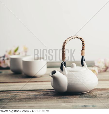 White Teapot, Cups, Sakura Flowers On Wooden Table Against The White Wall