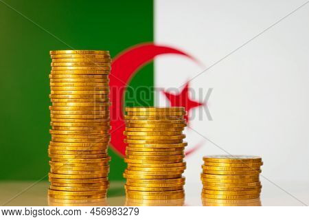 Three Stacks Of Golden Coins Forming Downward Graph On The Background Of Algeria Flag. Financial Cri