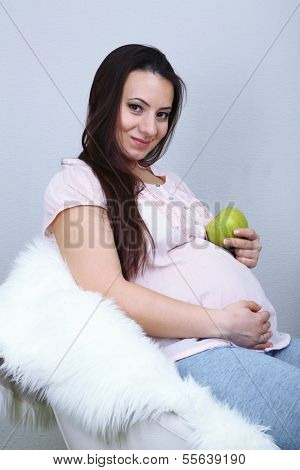 Young regnant woman sitting on armchair and holding apple on wall background