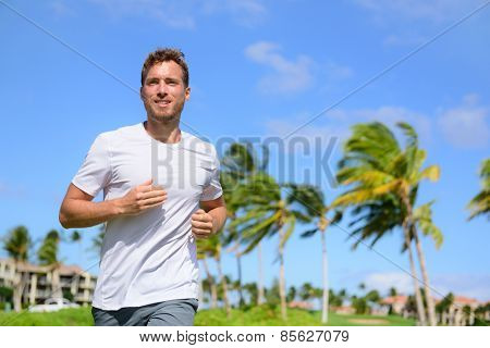 Healthy active man runner running in tropical park. Portrait of handsome young male jogger training cardio going for a run in city park or resort with palm trees in the background in summer.