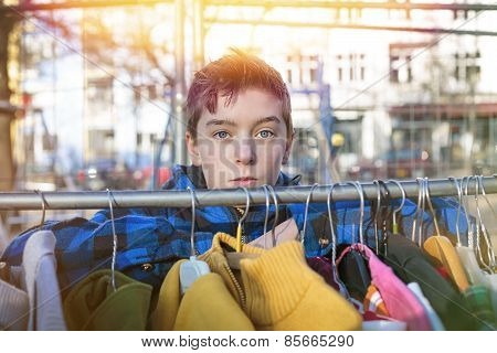 Portrait Of A Teenage Boy Behind A Clothes Rail On A Flea Market
