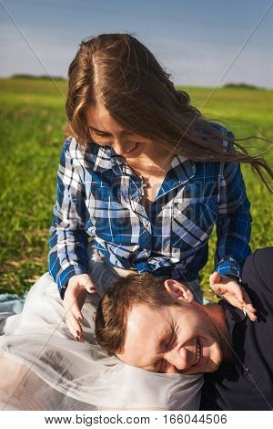 young girl in checkered shirt sitting on the grass. boy put his head on the girl's knees. They are happy and laughing