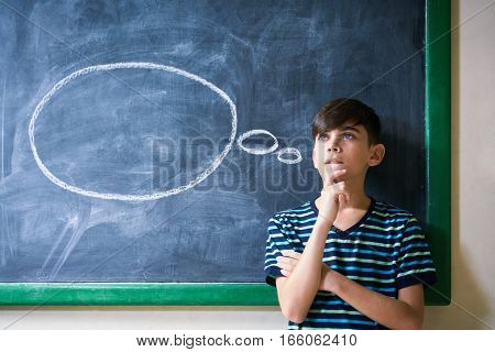 Concepts on blackboard at school. Intelligent and smart hispanic boy in class. Portrait of male kid thinking with hand on chin looking up against cloud drawing on blackboard