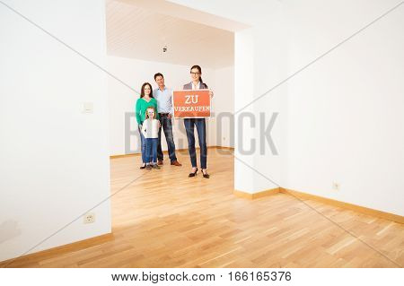 realtor and family in empty apartment, holding 'zu verkaufen' for sale sign