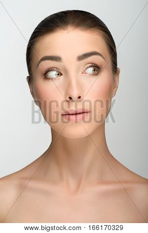 Young girl with big eyes and dark eyebrows, with naked shoulders, looking towards, a model with light nude make-up, gray studio background, beauty photo, close up.