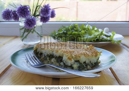 Portion of casserole with potatoes and spinach on a wooden table near the window. Jar with of chives flowers and plate with lettuce and parsley is on the table as well.
