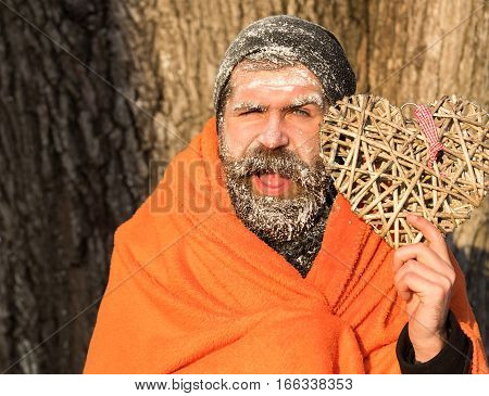 Frozen smiling man with squinty eyes bearded hipster with beard and moustache covered with white frost wrapped in orange blanket with wicker heart on winter day outdoors on natural background