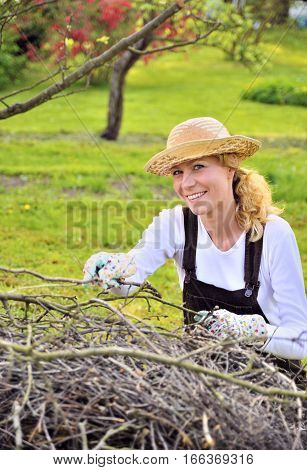Young woman working in orchard, after tree pruning, pile of cut branches and twigs of fruit trees, cutting branches of apple trees in garden, smiling woman piling up brushwood in spring