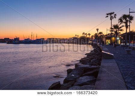 View over the seafront of the city Paphos in Cyrpus. Palm silhouettes, boats in the water and golden sunset over the sea with dramatic cloudy sky.