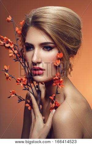Beautiful young woman with make-up and loose hairdo holding artificial sakura branch with orange flowers. Beauty shot on brown background. Copy space.