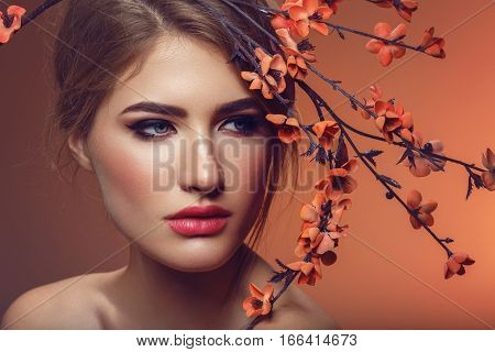 Beautiful young woman with make-up and loose hairdo with artificial sakura branch with orange flowers. Beauty shot on brown background. Copy space.