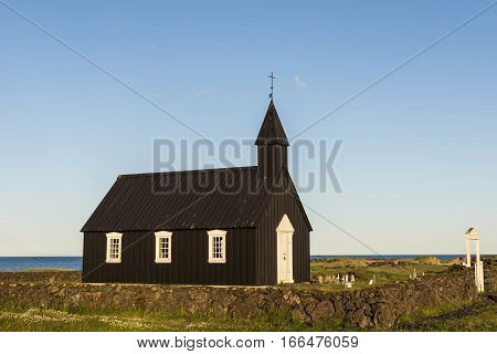 Black wooden Budakirkja at Saefellsnes with gate and stone wall on Iceland in evening light.
